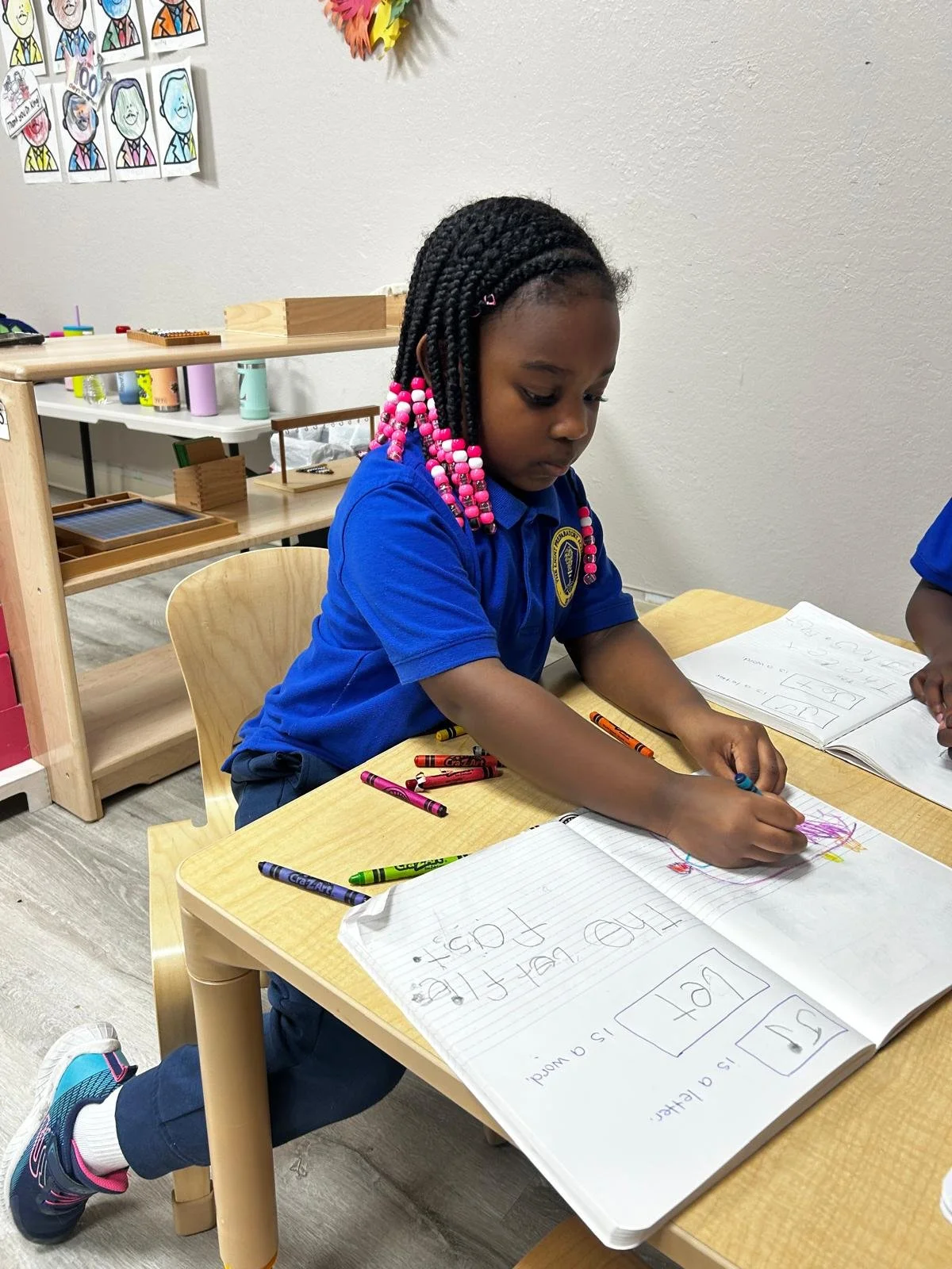 A young girl in a blue school uniform with pink and white beads in her hair, sitting at a wooden table and coloring in a notebook. Classroom artwork is on the wall behind her.