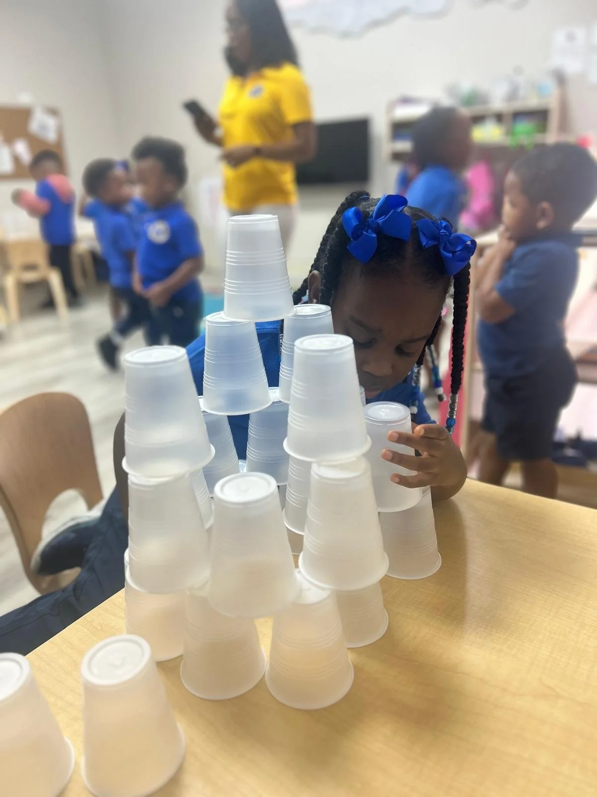 A young girl with braided hair and blue ribbons plays with stacked plastic cups on a table in a classroom with children and a teacher in the background.