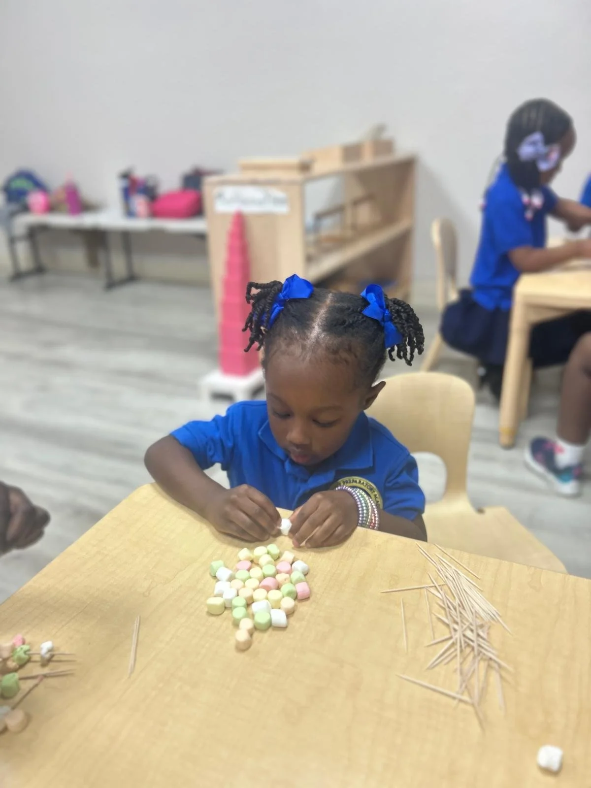 Young girl with braids tied with blue bows, sitting at a wooden table, counting small, colorful marshmallows with toothpicks nearby, in a classroom setting.