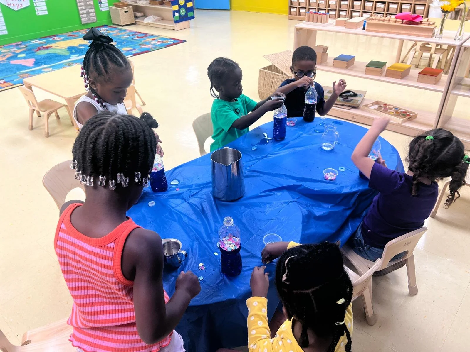 Six young children sitting and standing around a table in a classroom, conducting a science activity with bottles filled with colored liquids and small containers, engaging in hands-on experiments.