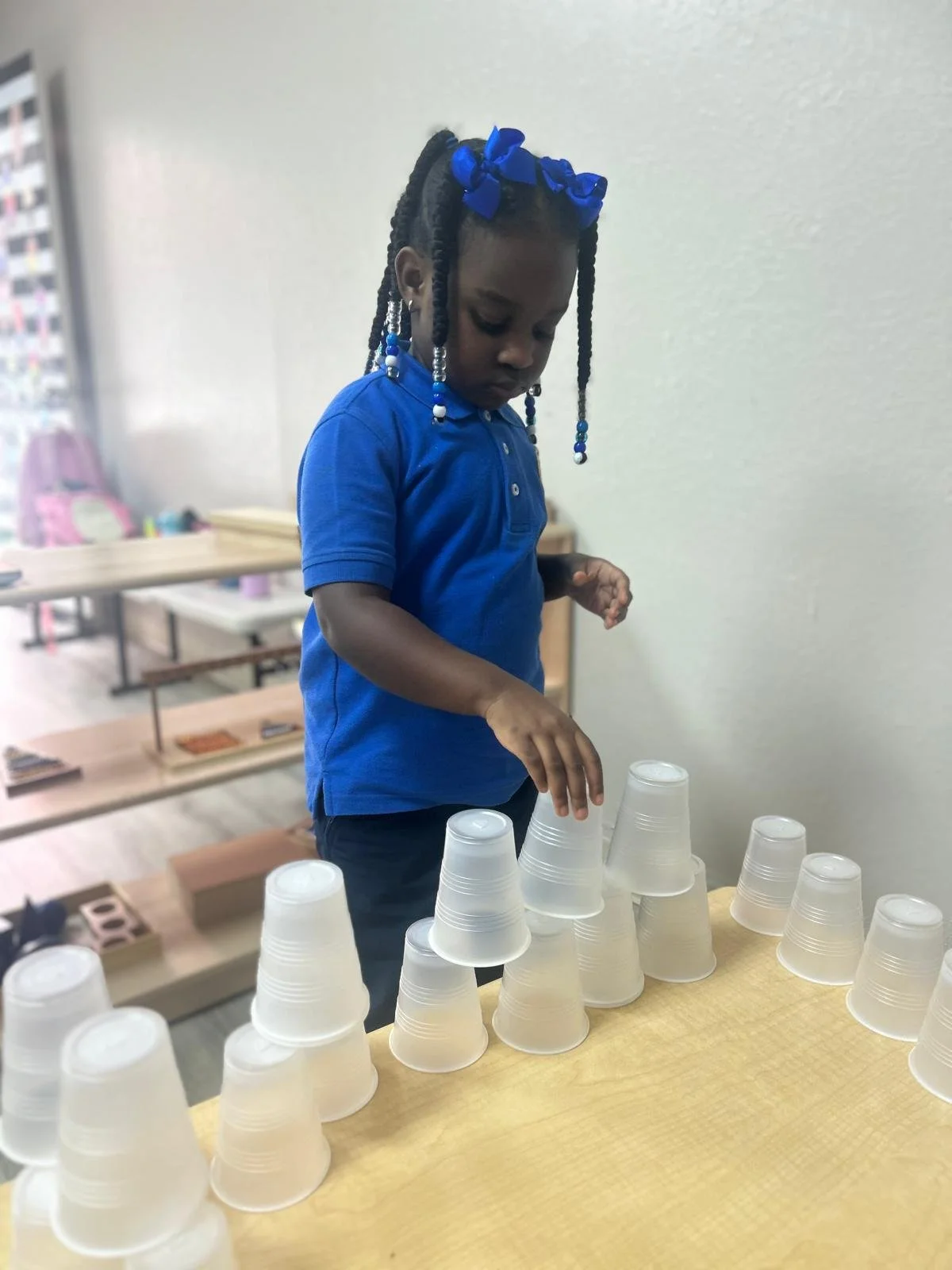 A young girl with braided hair and a blue bow wearing a blue shirt stacking clear plastic cups in a classroom or playroom.