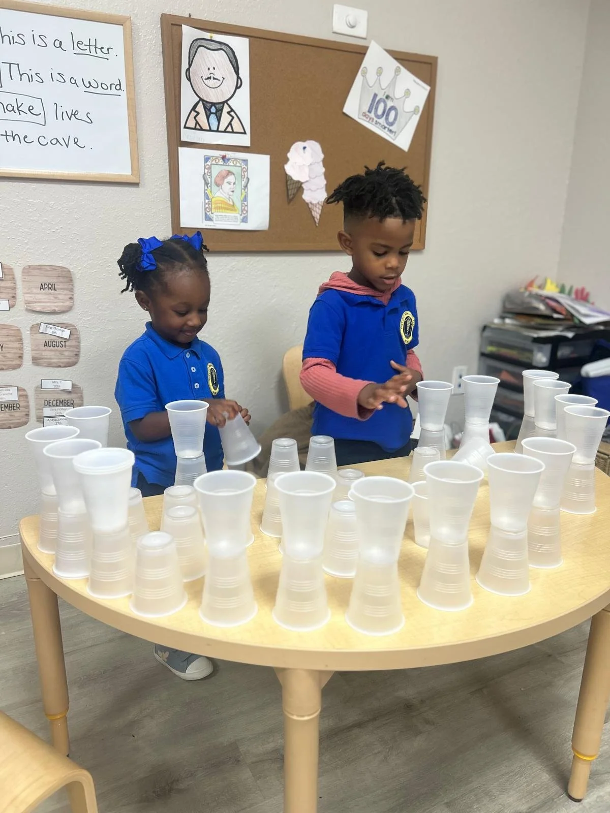 Two young children, a girl and a boy, wearing blue school uniforms, are standing at a light-colored wooden table stacking clear plastic cups. The girl has dark hair styled with blue hair bows. The boy has dark, curly hair. Behind them is a classroom wall with educational posters and cardboard panels labeled with months of the year.