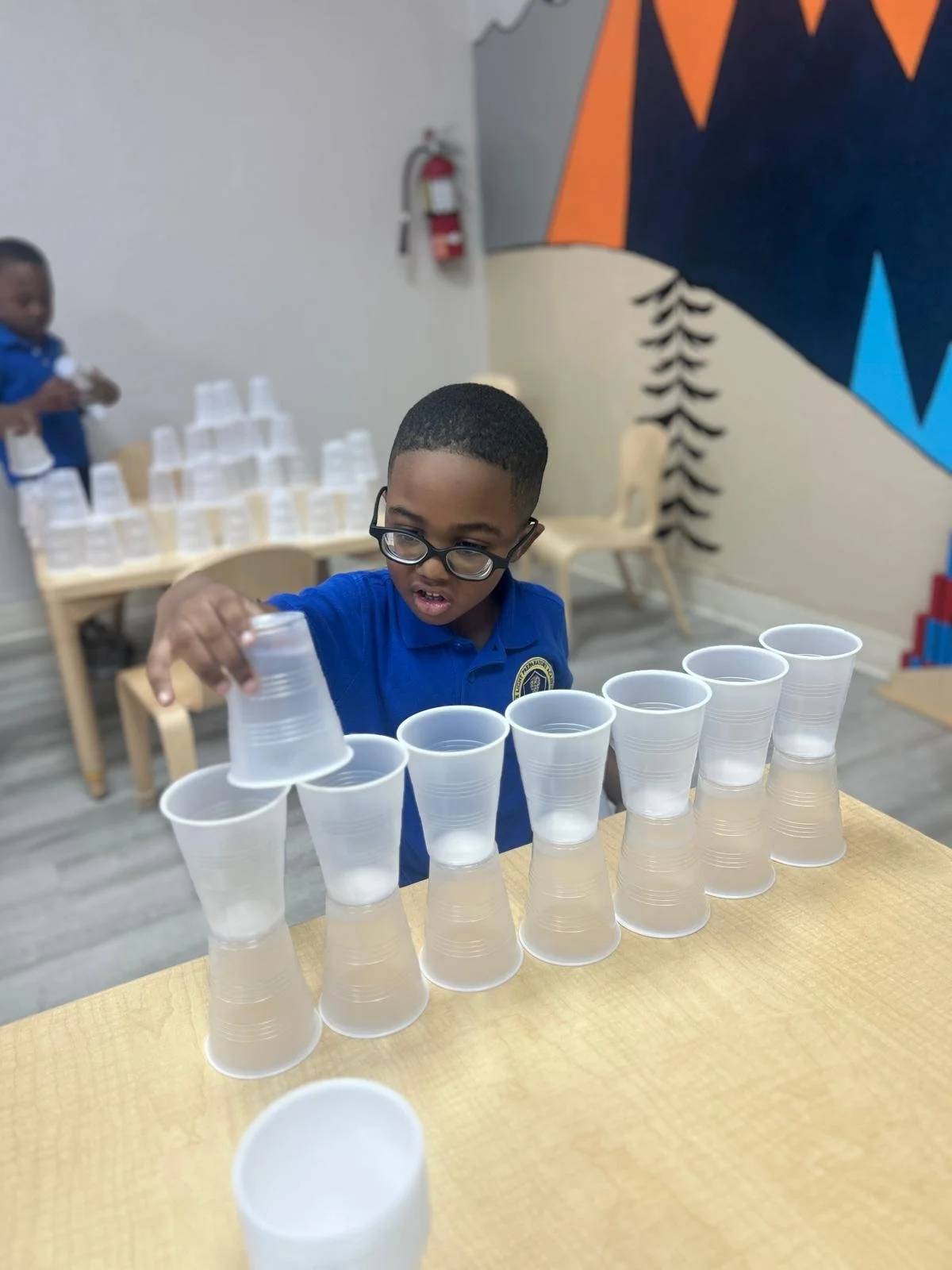 A young boy wearing glasses and a blue shirt stacking plastic cups on a table, with a mural and a fire extinguisher in the background.