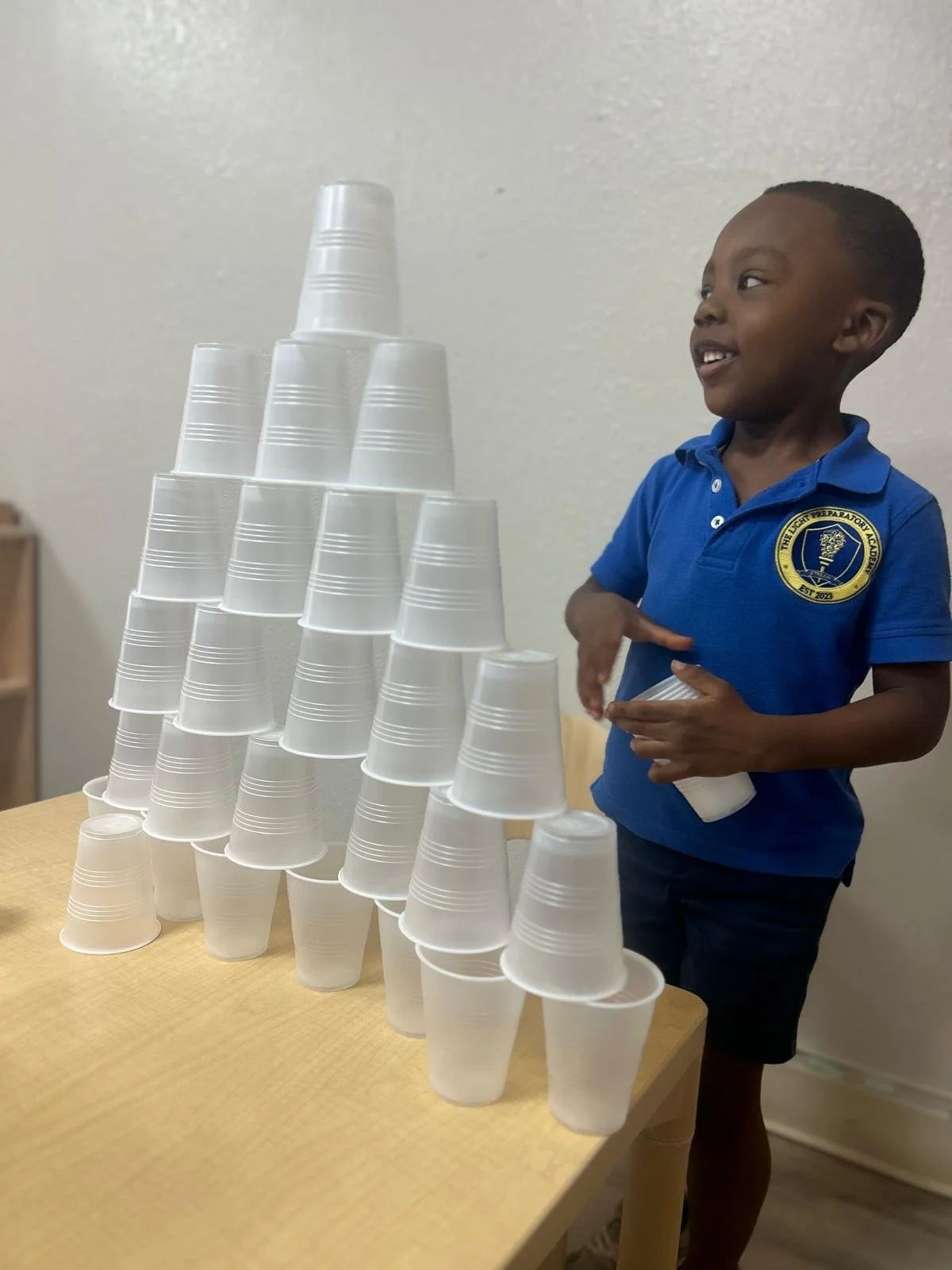 A young boy in a blue shirt smiling as he stacks white disposable cups into a pyramid on a wooden table.