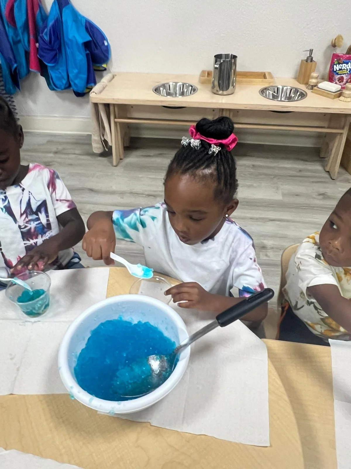 Children playing with blue jelly-like substance at a table in a classroom.