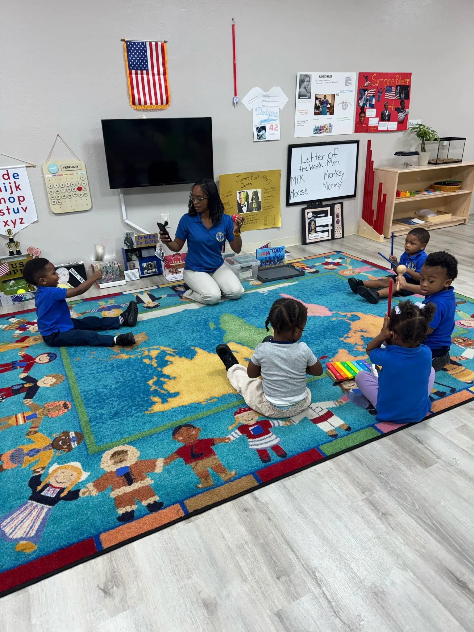 A teacher with glasses and braids sitting on a colorful rug with children in a classroom, teaching and playing mounted on the wall are an American flag, whiteboard with words, educational posters, and a TV.