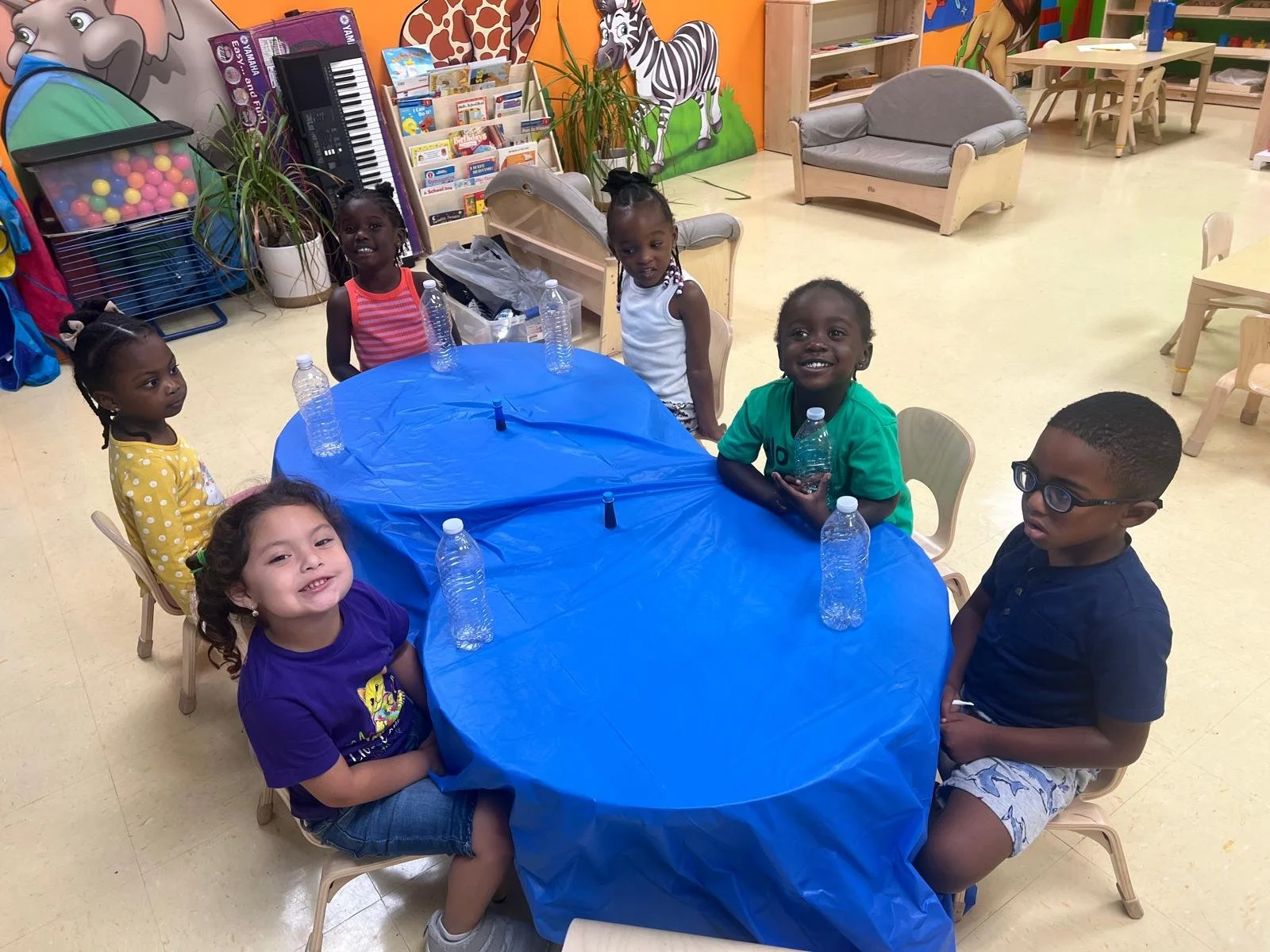 Children sitting around a table with a blue cover, holding water bottles, in a colorful indoor playroom or classroom.