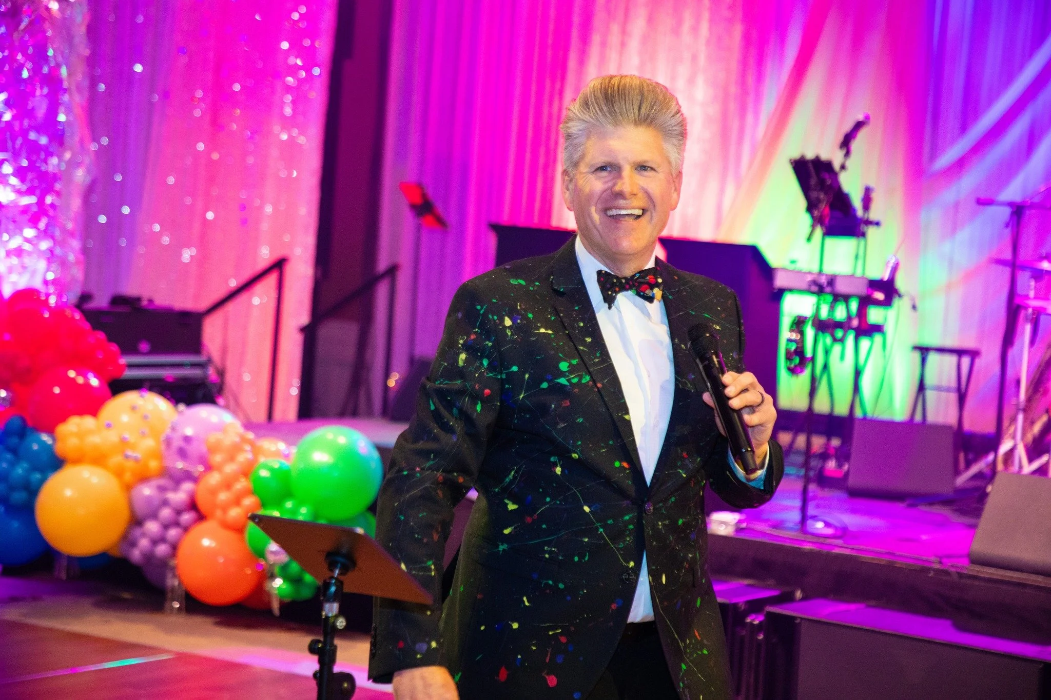 Bobby D., auctioneer, in a colorful suit and bow tie holding a microphone, smiling, standing in front of a vibrant, pink and purple brightly lit stage decorated with balloons.