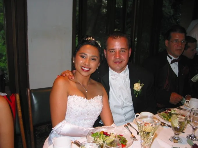 A bride and groom sitting at a wedding reception table, dressed in formal attire, with plates of food and glasses on the table.