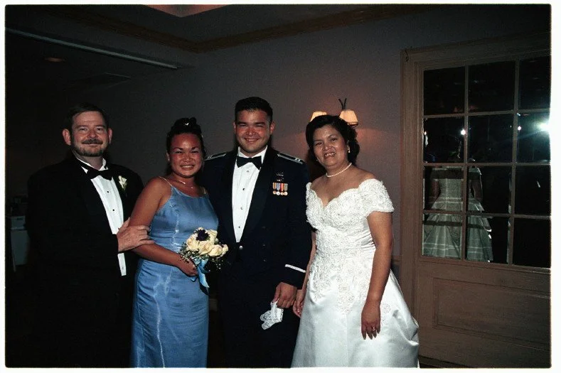 Group photo at a wedding with four people in formal attire. A man in a tuxedo stands next to a woman in a blue dress holding a bouquet. Another man in a military uniform stands beside a woman in a white wedding dress. A mirror in the background refle
