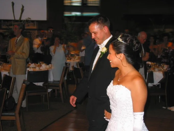 Bride and groom smiling at their wedding reception, walking together in formal attire with guests in the background applauding.