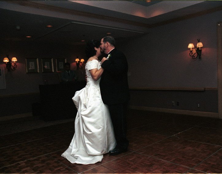 Bride and groom dancing in a dimly lit room, with wall sconces and framed pictures in the background.