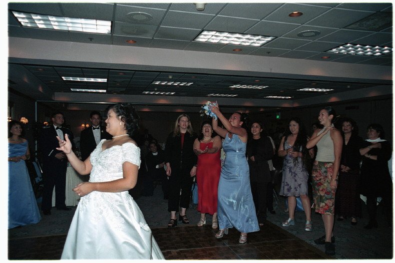 Bride throwing bouquet at a wedding reception, with several women reaching to catch it while other guests watch in the background.