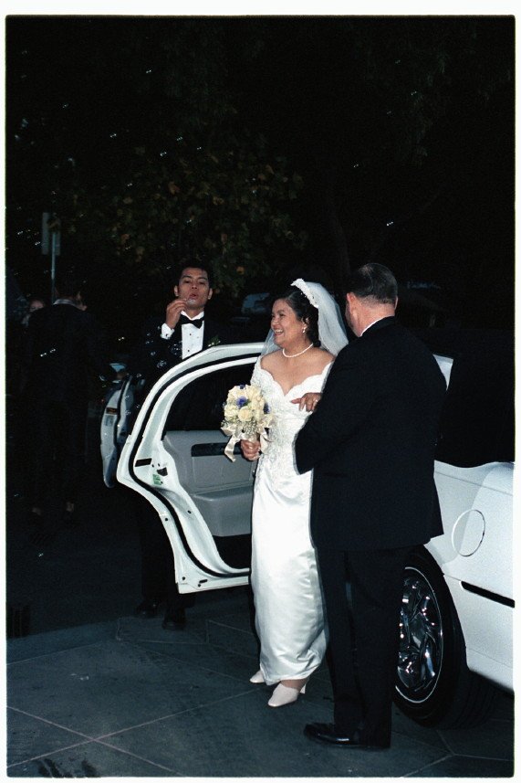 Bride in a white dress holding flowers, accompanied by two men in tuxedos, standing next to a white limousine with an open door at night.
