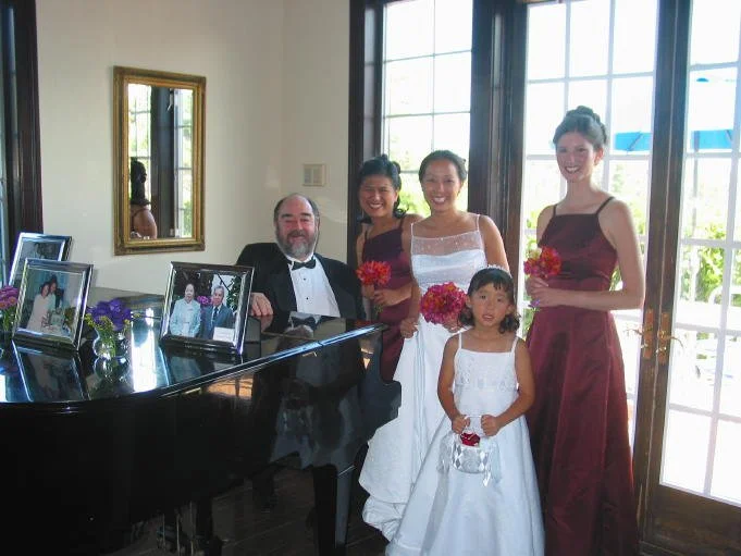 A man in a tuxedo sitting at a grand piano with framed photos on top, surrounded by three women in formal dresses and a young girl in a white dress, all holding flowers, inside a bright room with large windows.