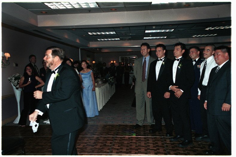 A man in a tuxedo tossing a small object, possibly a garter, towards a group of men at a formal event, likely a wedding reception. Guests dressed in formal attire are gathered in a hall with a dining table and floral decorations in the background.