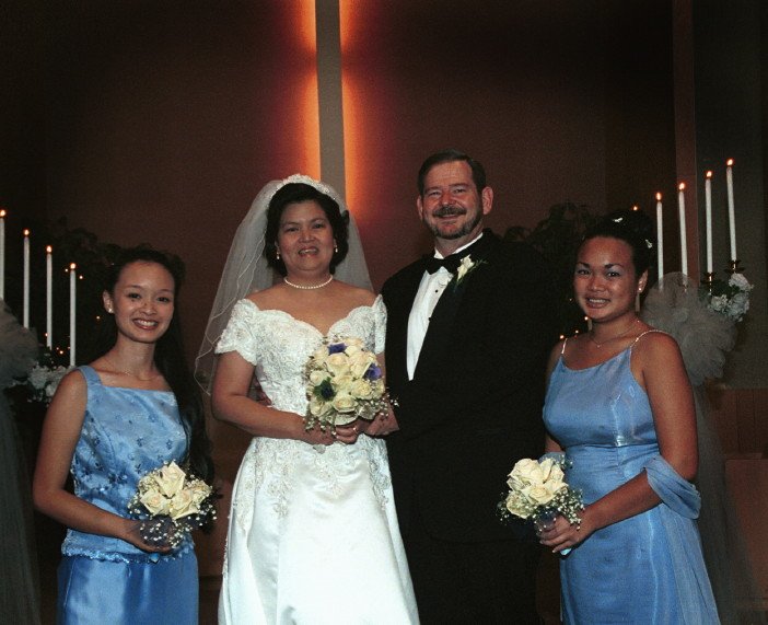 A bride and groom standing with two bridesmaids at a wedding ceremony. The bride is wearing a white dress with a veil, and the groom is in a black tuxedo. The bridesmaids are dressed in light blue gowns and holding bouquets. The background shows lit 