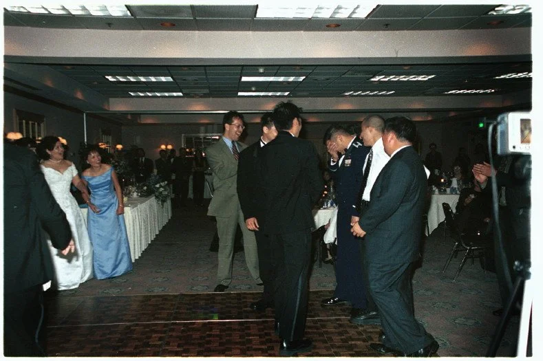 People dressed formally at a wedding reception, with men in suits and women in gowns; some are dancing or interacting while others are seated in a banquet hall.