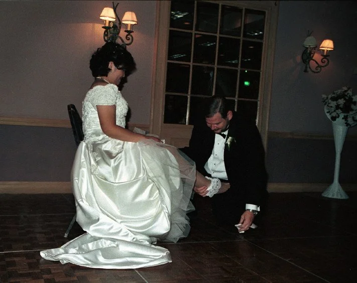 A bride in a white wedding dress sits on a chair while a groom in a tuxedo kneels on the floor, attempting to remove the garter from her leg, in a dimly lit room with wall lights and a large window.