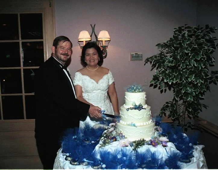 A couple in formal attire cutting a tiered wedding cake adorned with blue and purple decorations, standing in a decorated room with foliage and a wall sconce.