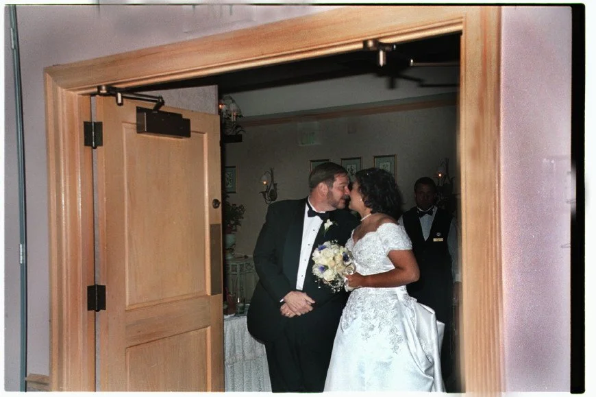 A bride and groom in formal attire are sharing a tender moment, standing in a doorway. The groom is wearing a black tuxedo with a bow tie, and the bride is in a white lace gown holding a bouquet of flowers.