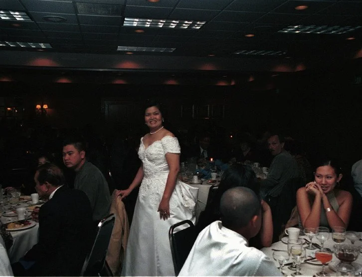 A bride in a white wedding dress stands smiling at a formal event, surrounded by seated guests at round tables in a dimly lit venue.