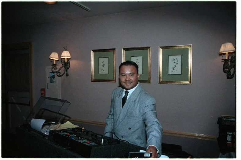 A man in a suit standing next to DJ equipment in a room with framed botanical pictures and wall sconces.