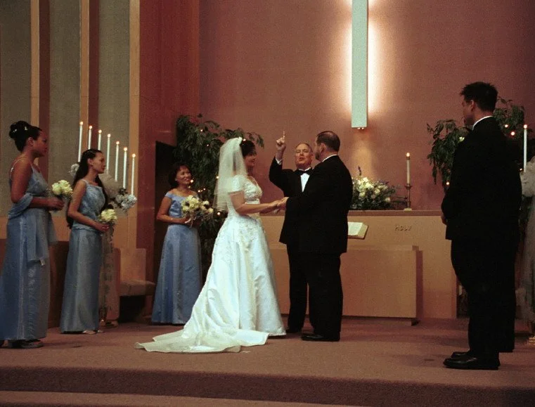 A wedding ceremony in a church with a bride in a white gown and veil holding hands with the groom, who is in a black suit. Bridesmaids in blue dresses stand to the left, a groomsman to the right. A minister stands behind the couple, beside an altar with candles and greenery.