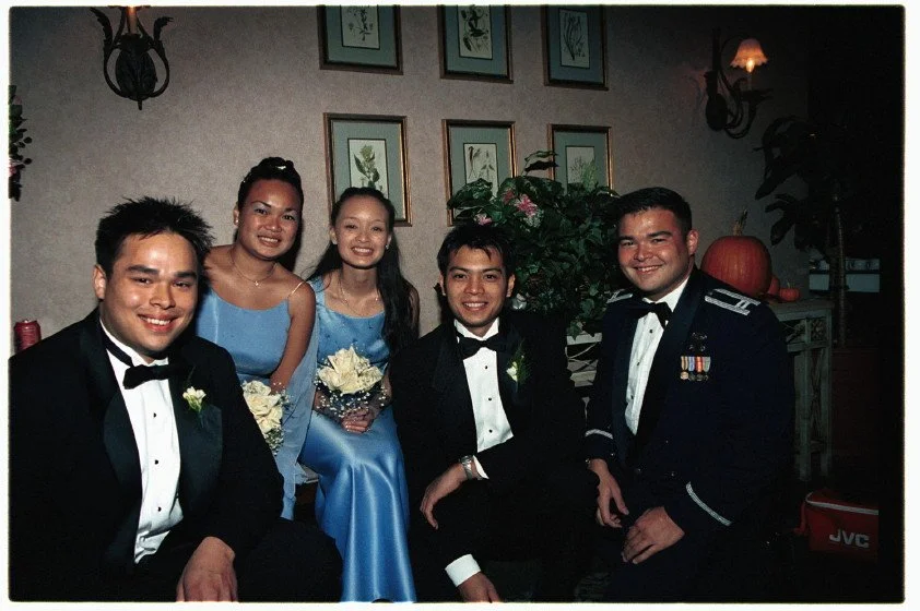 Group of five people in formal attire, including three men in tuxedos and two women in blue dresses, sitting together indoors. The setting has wall art and plants, and a pumpkin is visible on the right.