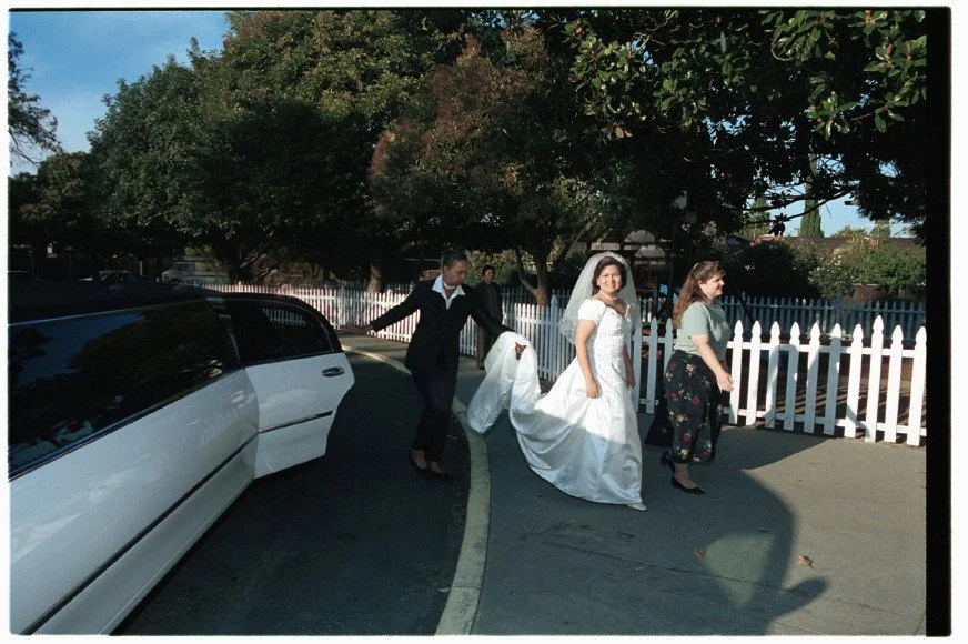 Bride in a white wedding dress walking with attendants near a limousine parked on a street, surrounded by trees and a white picket fence.
