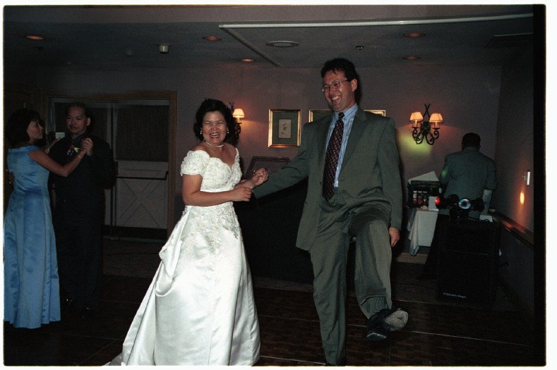 A bride and groom dancing at a wedding reception, both smiling joyfully. The bride is wearing a white gown, and the groom is in a suit. Other guests are also dancing in the background.