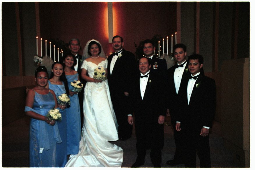 Wedding party group photo with bride, groom, bridesmaids in blue dresses, and groomsmen in tuxedos inside a church with lit candles.