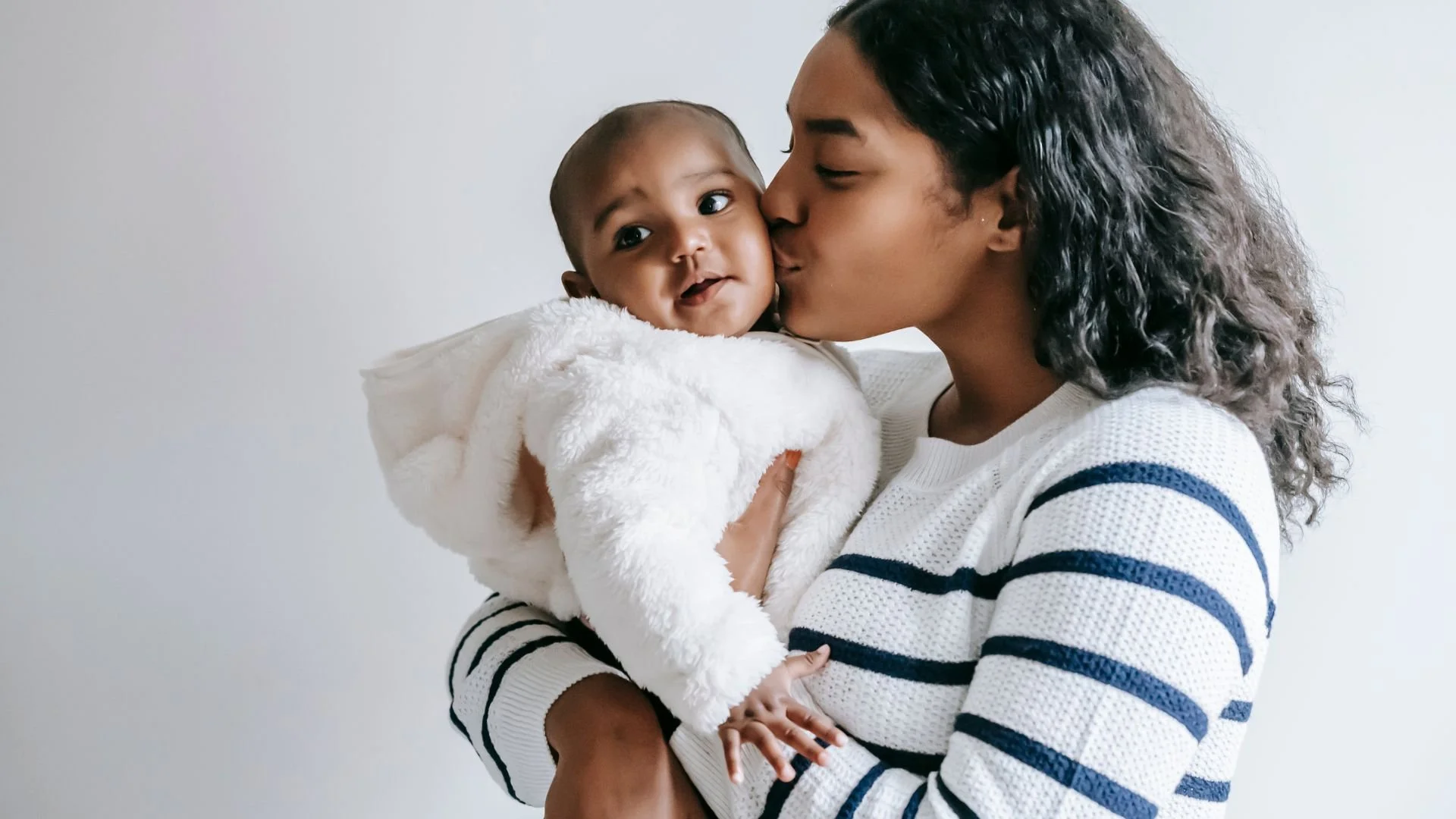 Woman kissing a baby wearing a fluffy white onesie in a tender moment.