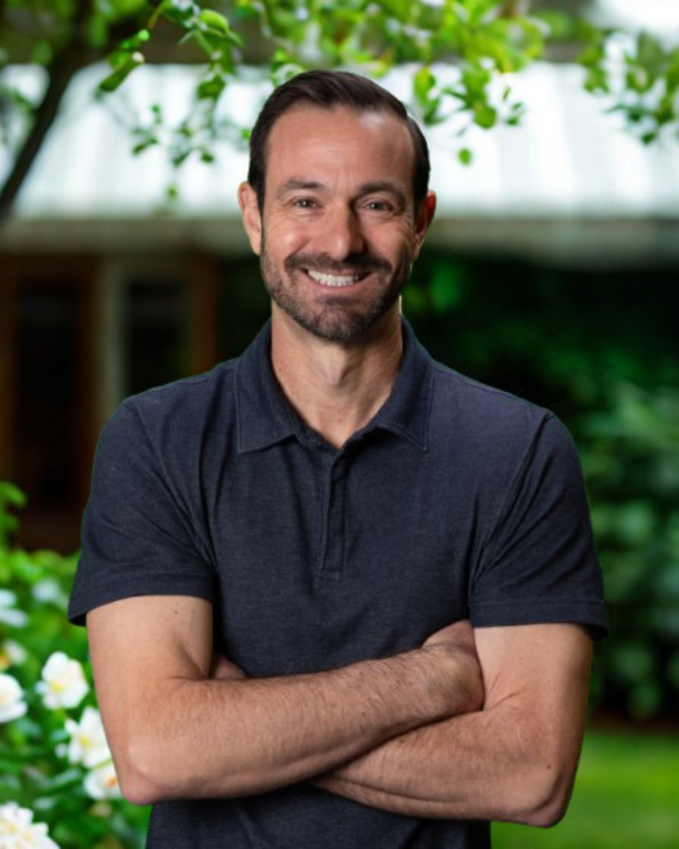 Man smiling wearing a navy golf shirt against a leafy background