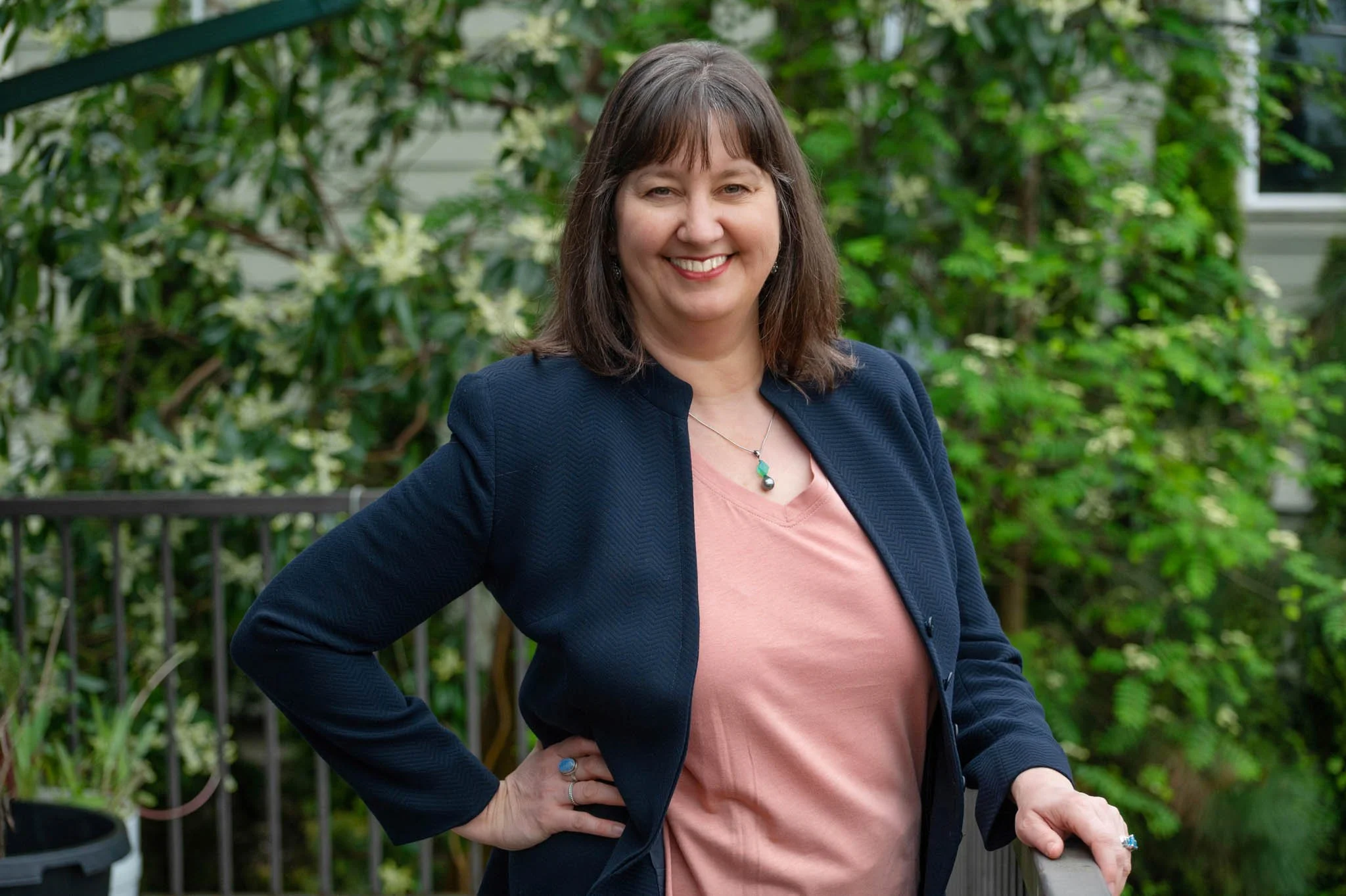 A woman smiling with a hand on her hip wearing a navy blazer and pink shirt against a leafy background