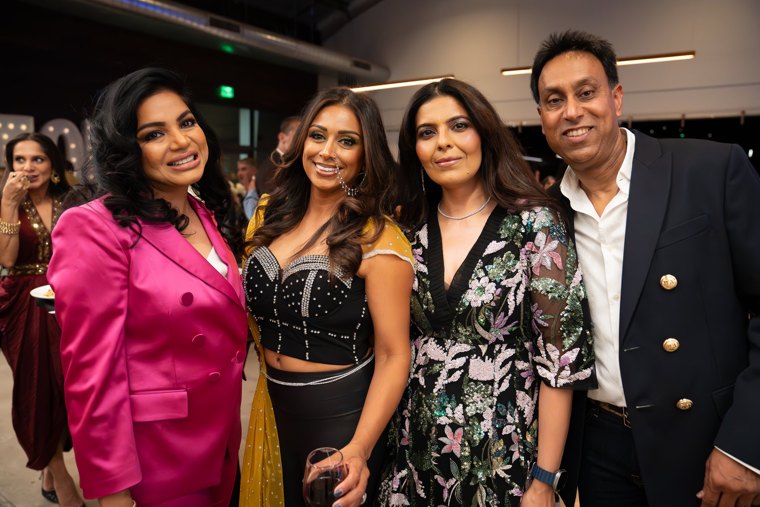 Group of four people posing, two women wearing colorful dresses, one in a pink blazer, one in a black and gold outfit, another in a floral dress, and a man in a suit. They are indoors, likely at an event.