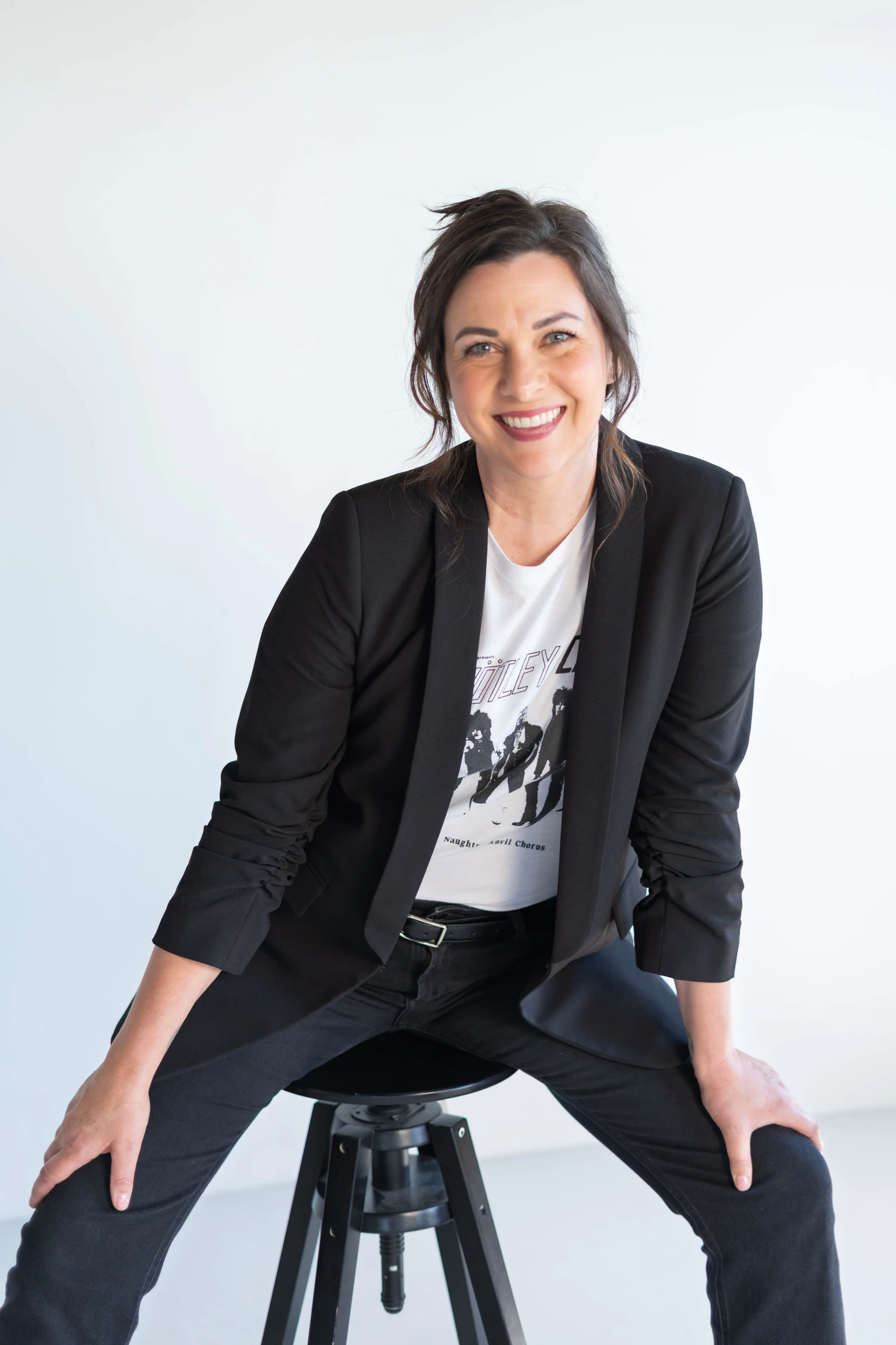 A woman with dark brown hair and light skin is sitting on a black stool, smiling, in a black blazer, graphic t-shirt, and black pants against a plain white wall.