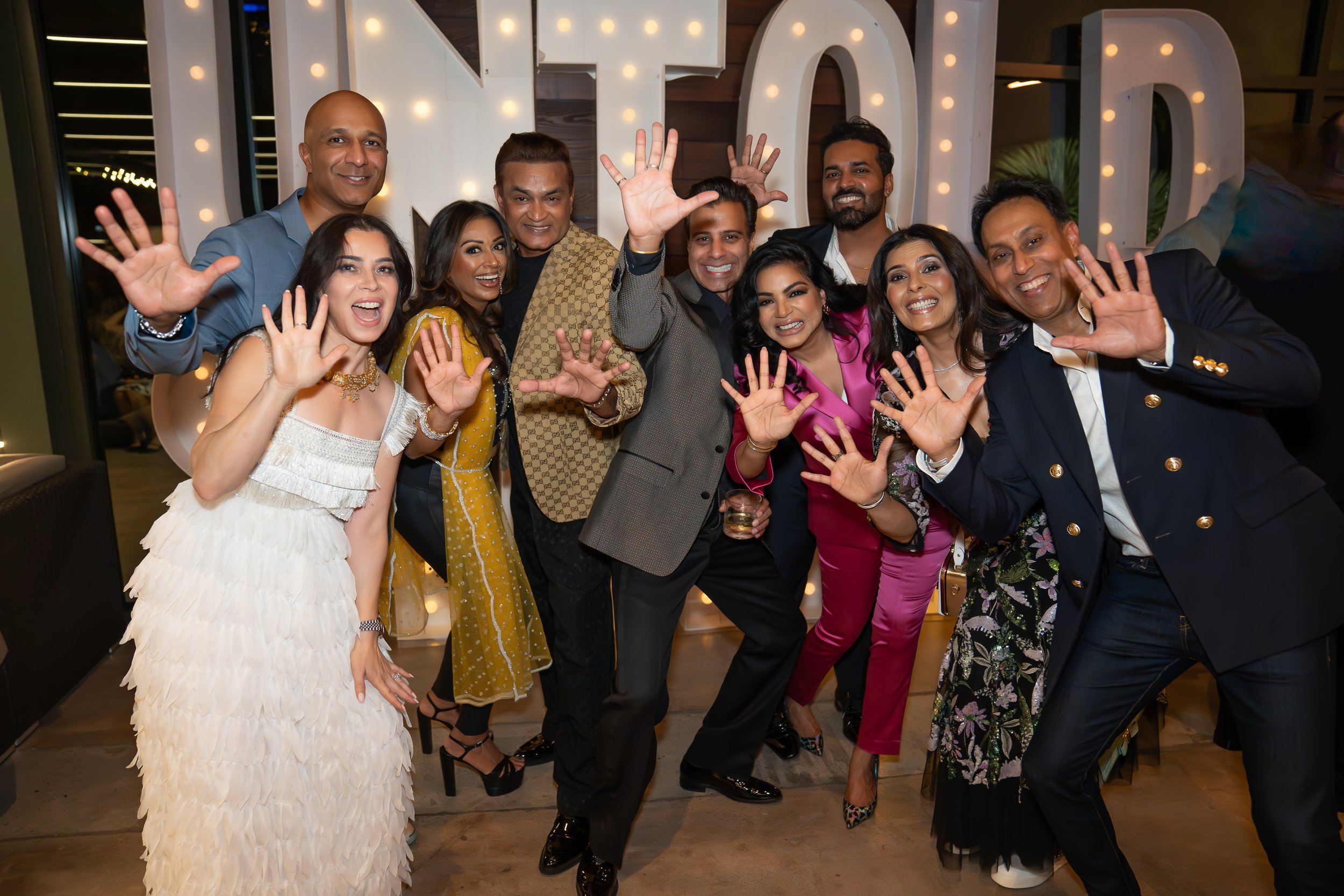 Group of nine people smiling and posing playfully in front of illuminated letters holding drinks.