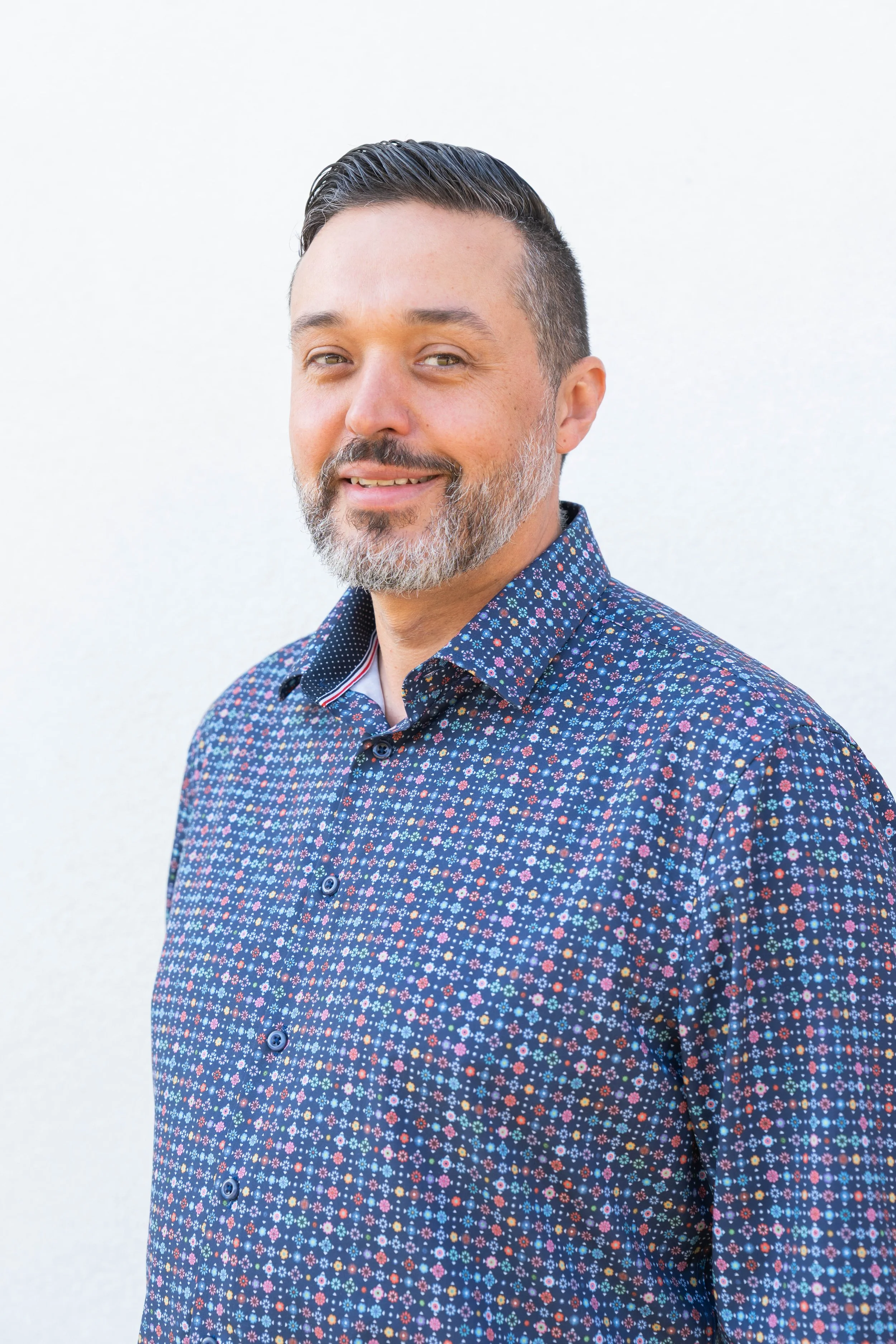Portrait of a middle-aged man with a beard and short dark hair, wearing a blue patterned shirt, standing against a white background.