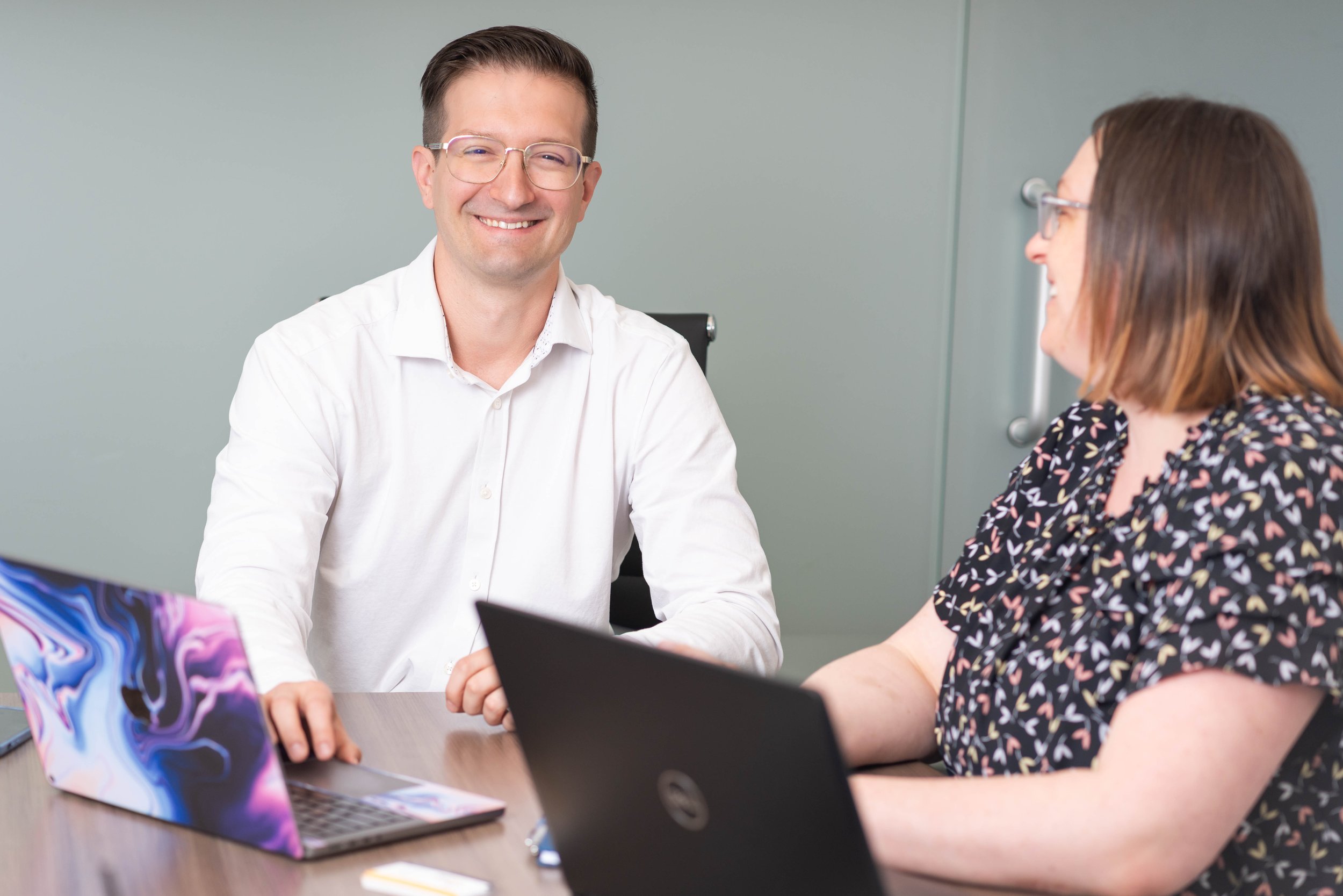 Two people sitting at a table with laptops, smiling, having a discussion in an office setting.