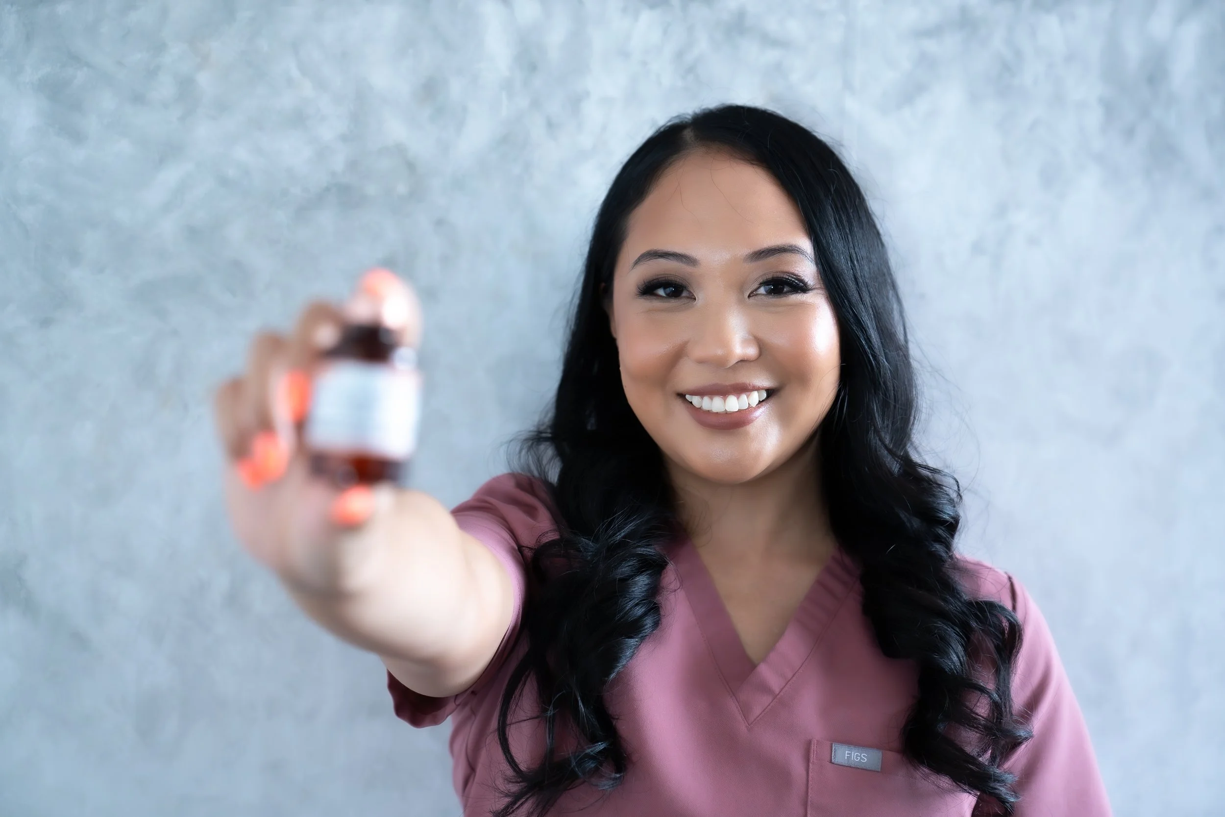 A smiling woman in pink scrubs holding a medication bottle towards the camera.