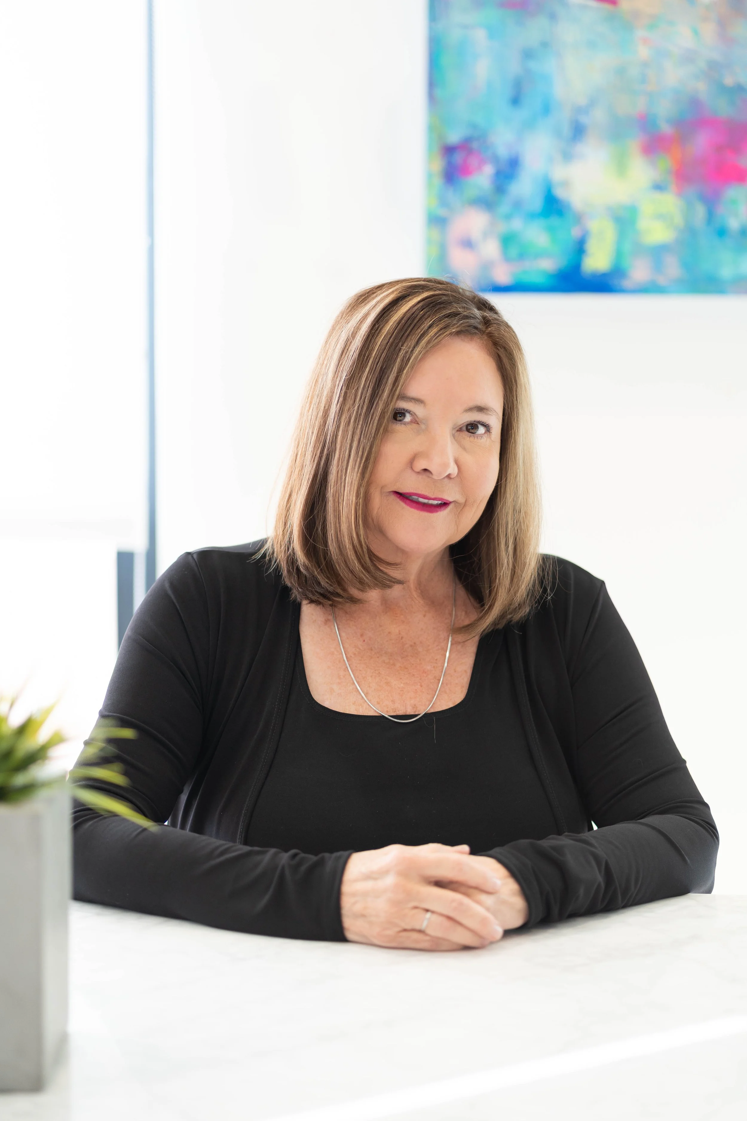 A woman with shoulder-length light brown hair, wearing a black top and a silver necklace, sitting at a white table in a bright room with an abstract painting on the wall behind her.