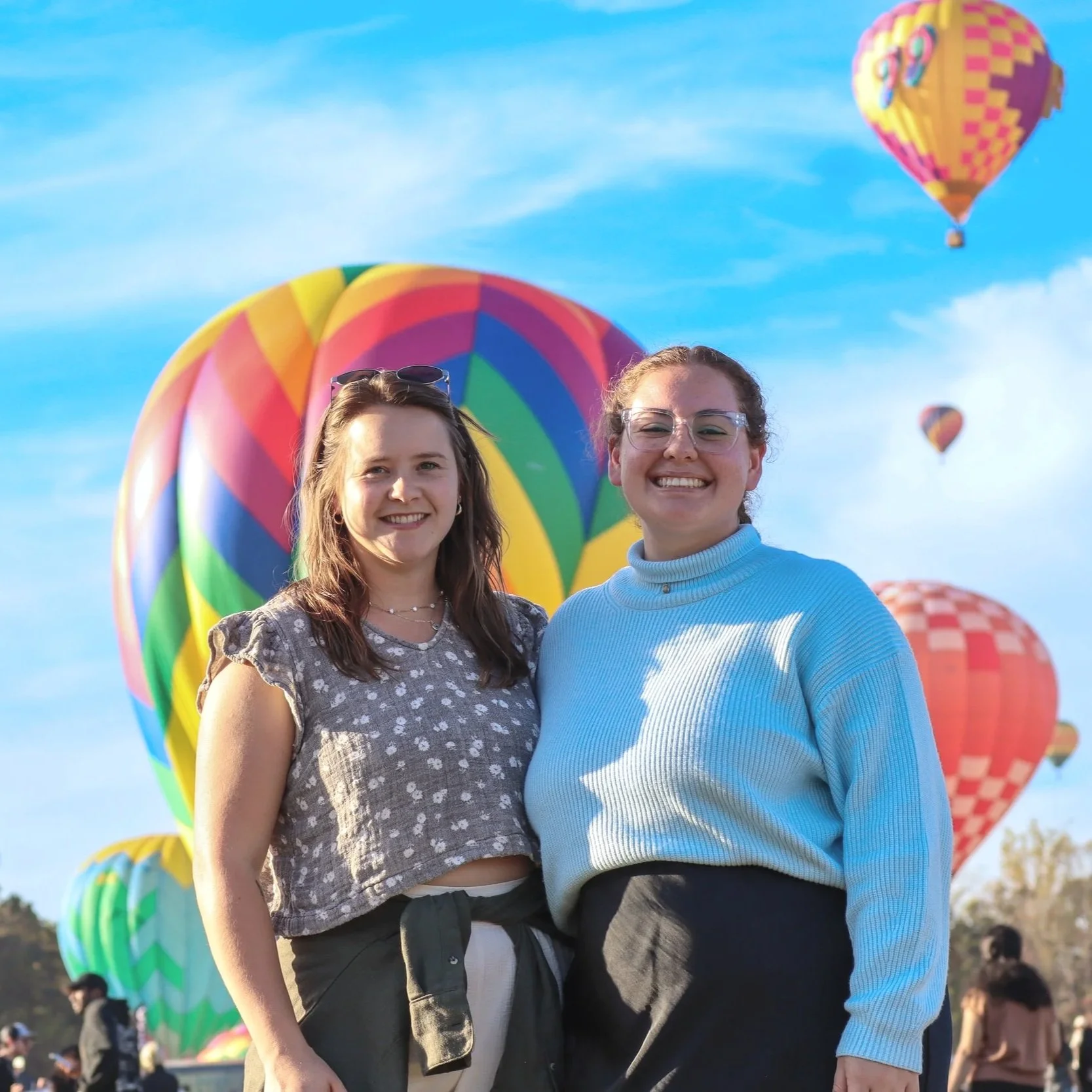 Two smiling young women standing in front of colorful hot air balloons in the sky, outdoors during daytime.