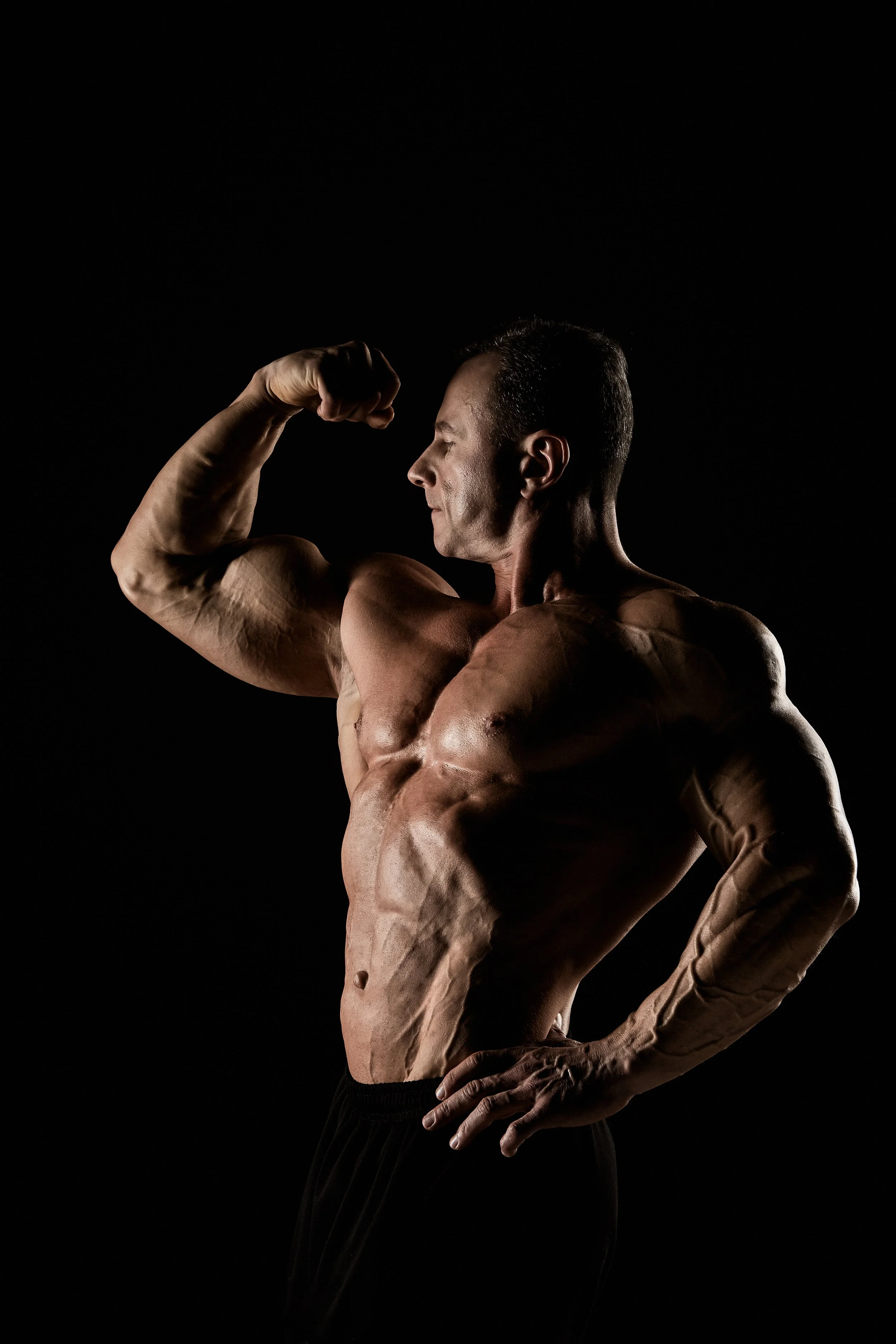 A muscular man flexing his bicep in a side pose, with a dark background highlighting his physique.