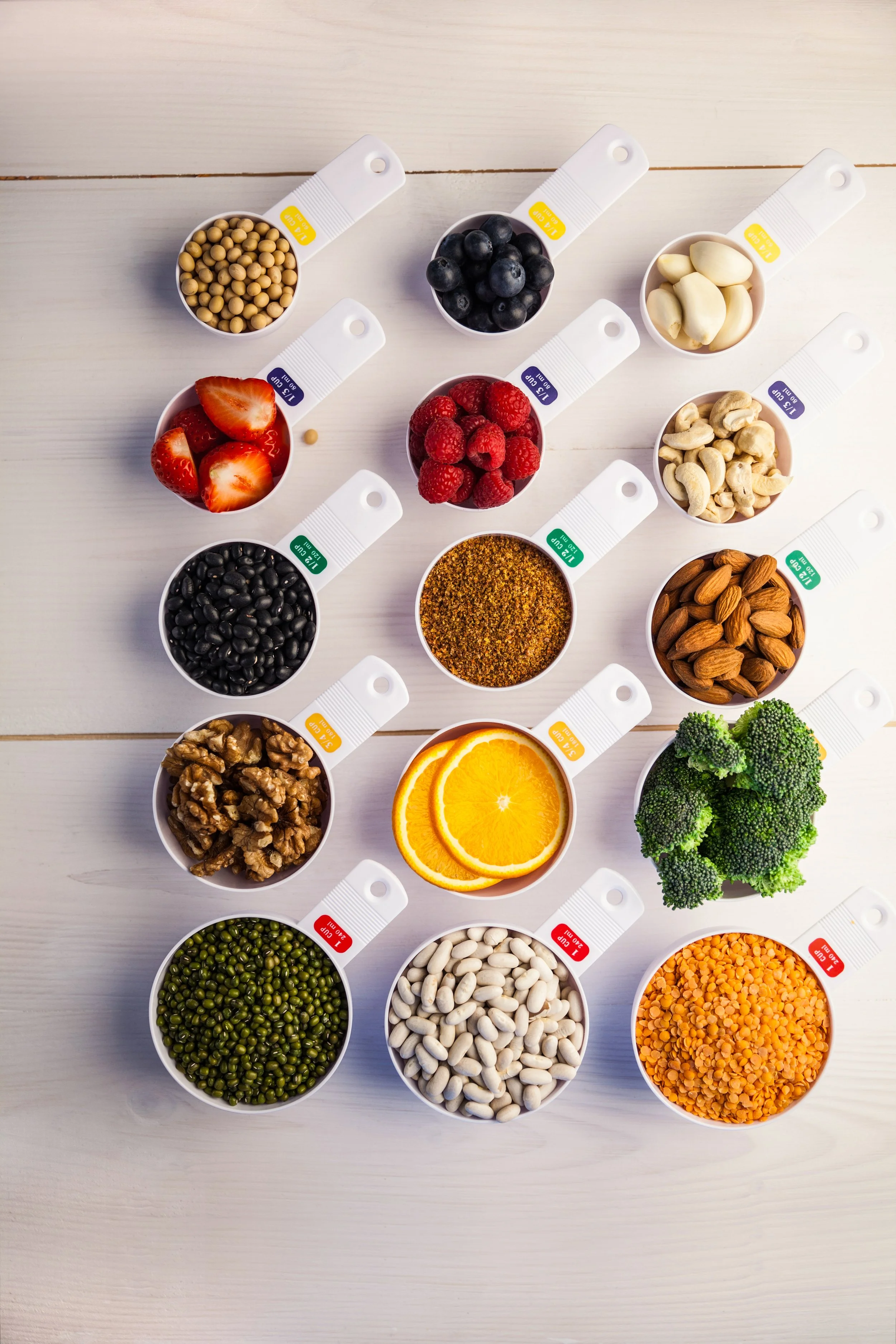 A variety of colorful food items in small white bowls arranged in a grid on a white wooden surface, including mixed vegetables, berries, nuts, and grains.