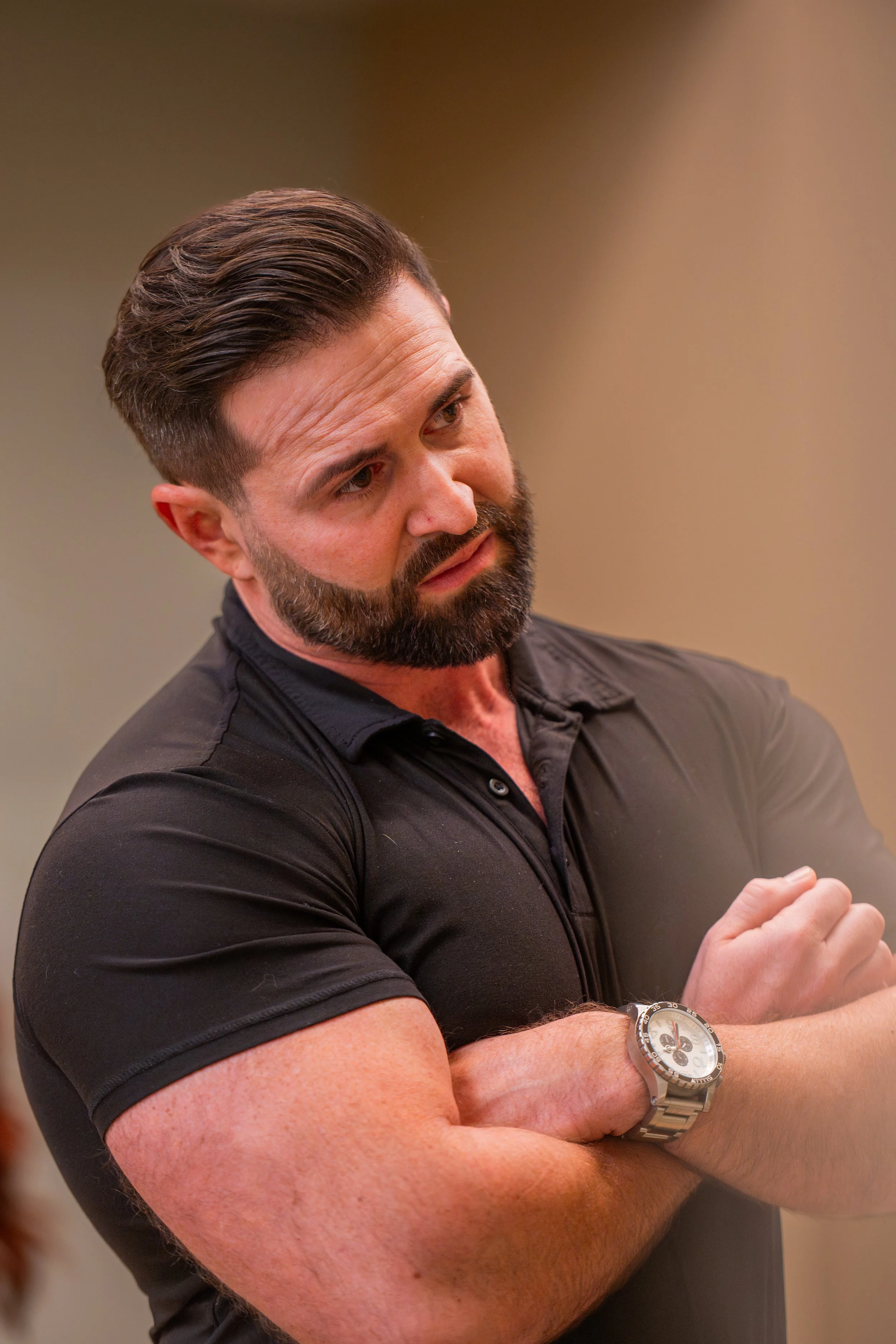 A man with a beard and dark hair, wearing a black polo shirt and a silver wristwatch, appears to be listening or contemplating, with arms crossed and a focused expression.