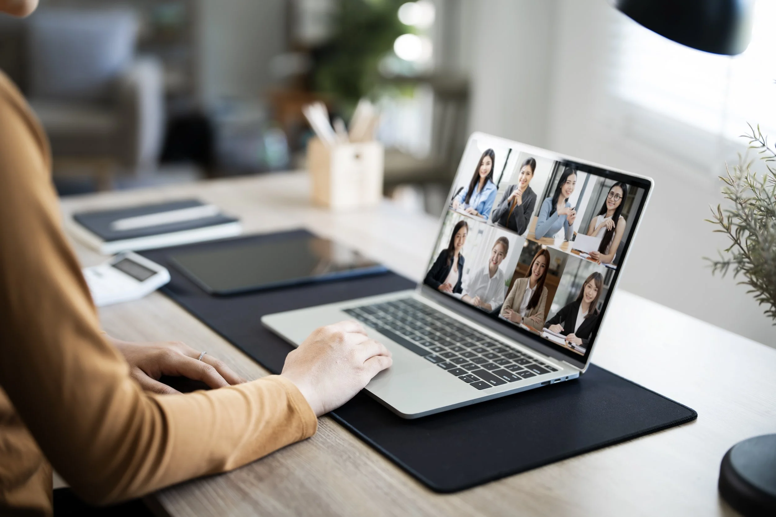 Person participating in a virtual video conference with multiple women on a laptop screen, seated at a desk with notebooks and tablets.
