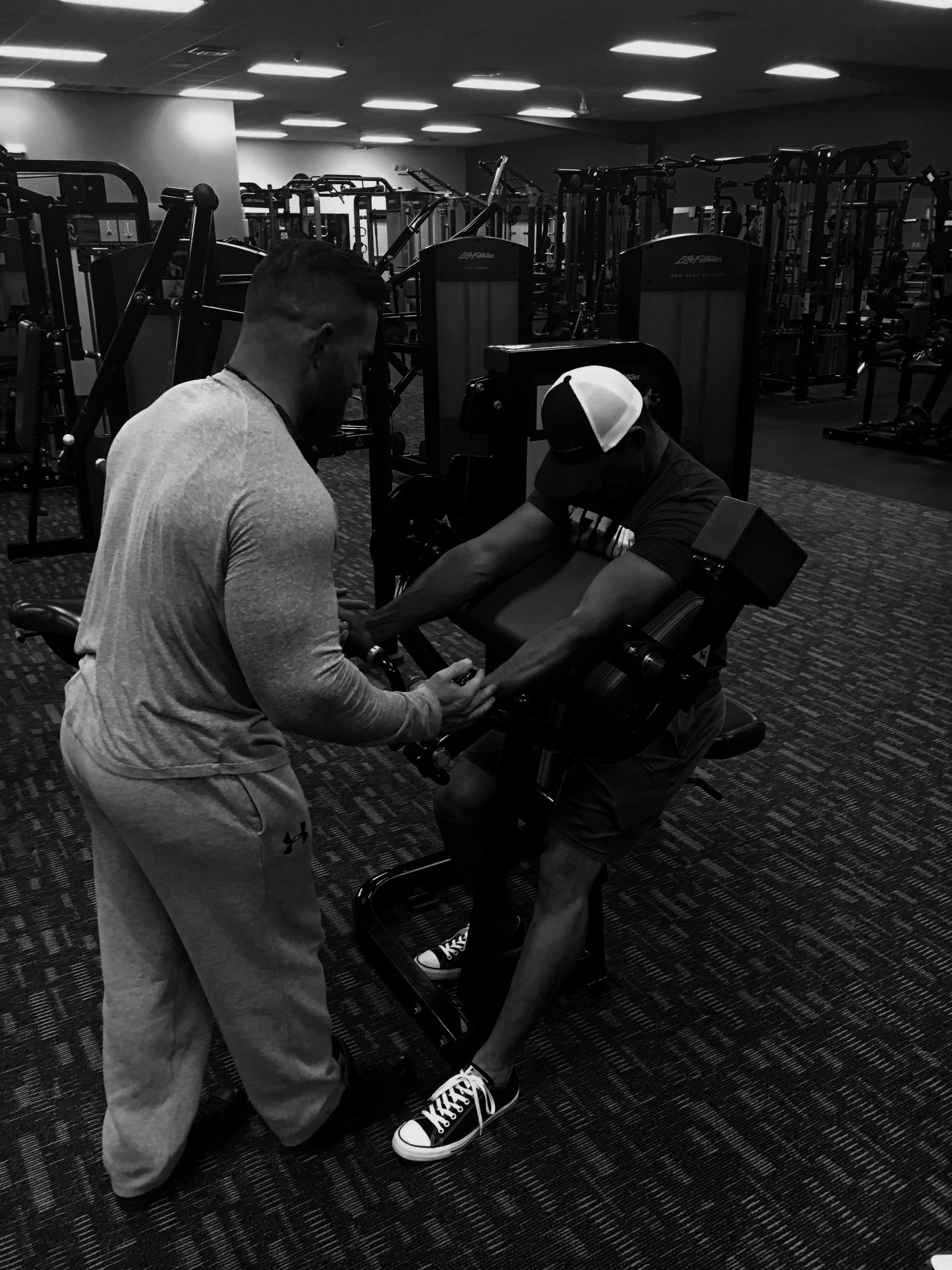 Two men, one spotting and assisting the other with strength training using a leg extension machine in a gym.