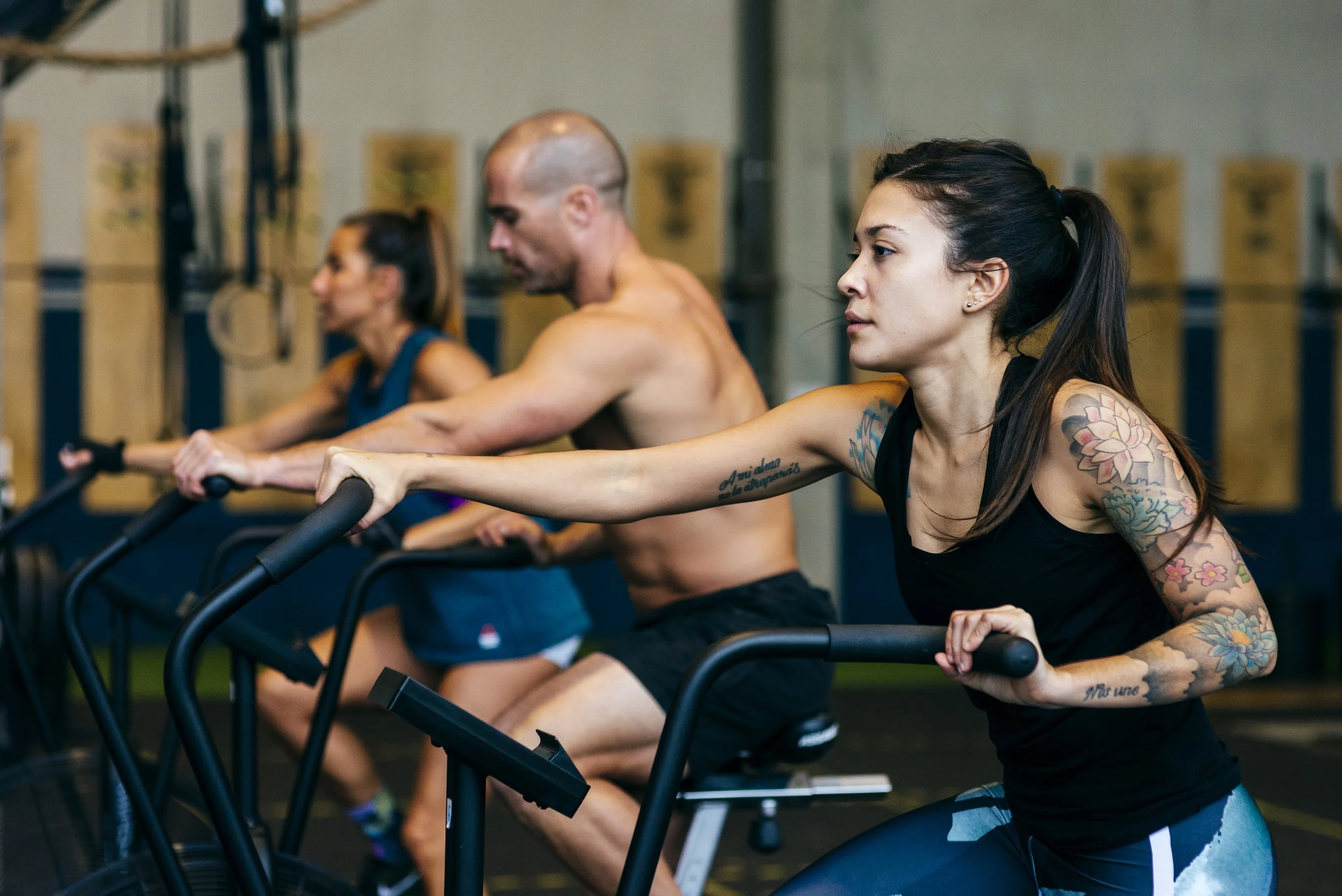 Three people using rowing machines in a gym, focusing on a woman with long dark hair and tattoos on her arms in the foreground, with two other individuals blurred in the background.