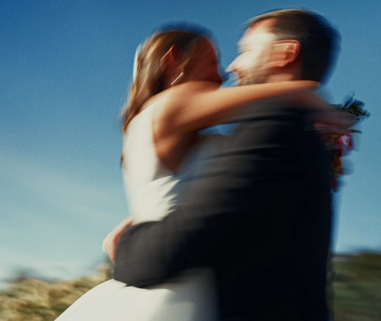 A blurred photograph of a couple embracing outdoors against a clear blue sky.